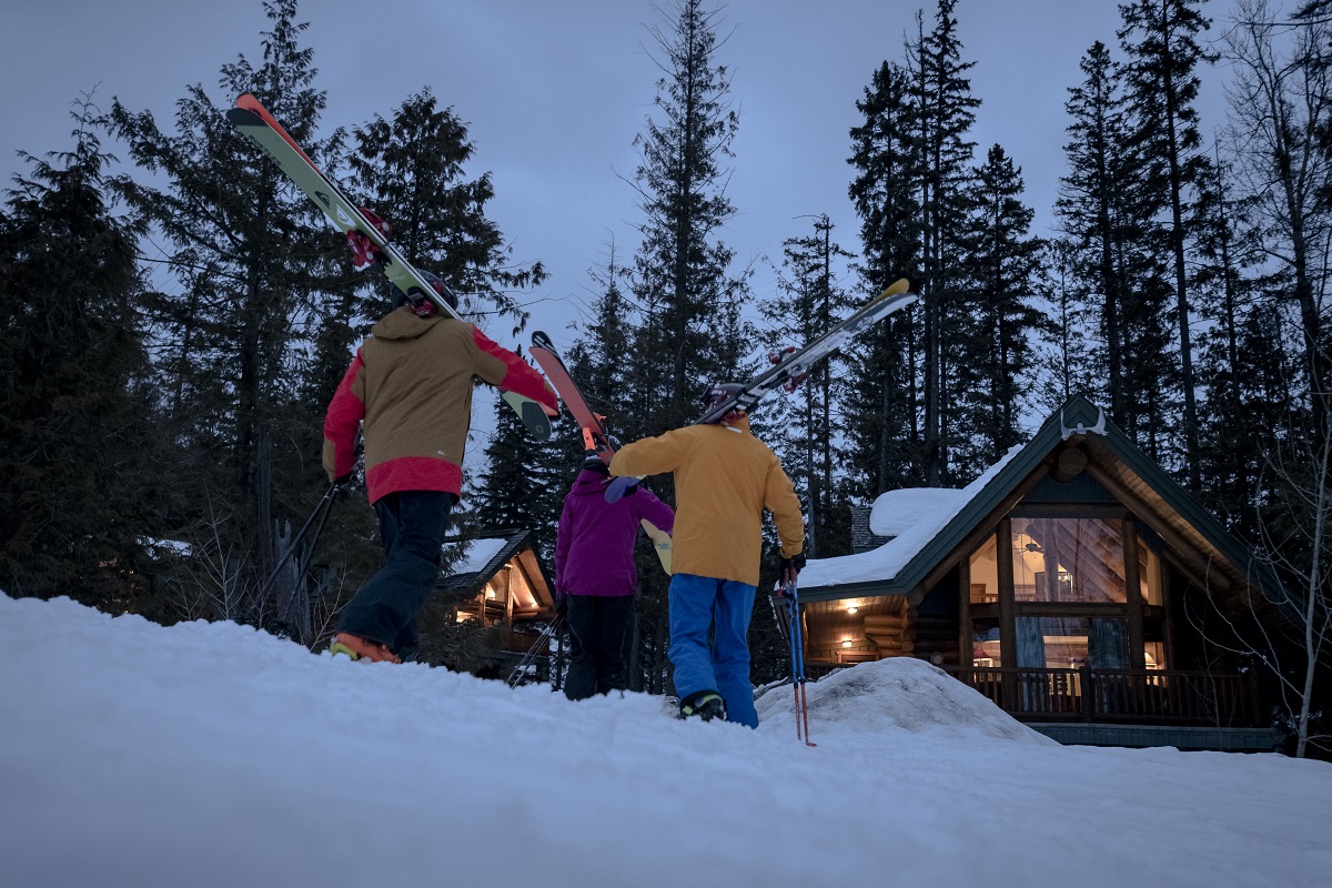 A group of skiers walk back to a mountain lodge in the evening carrying their skis