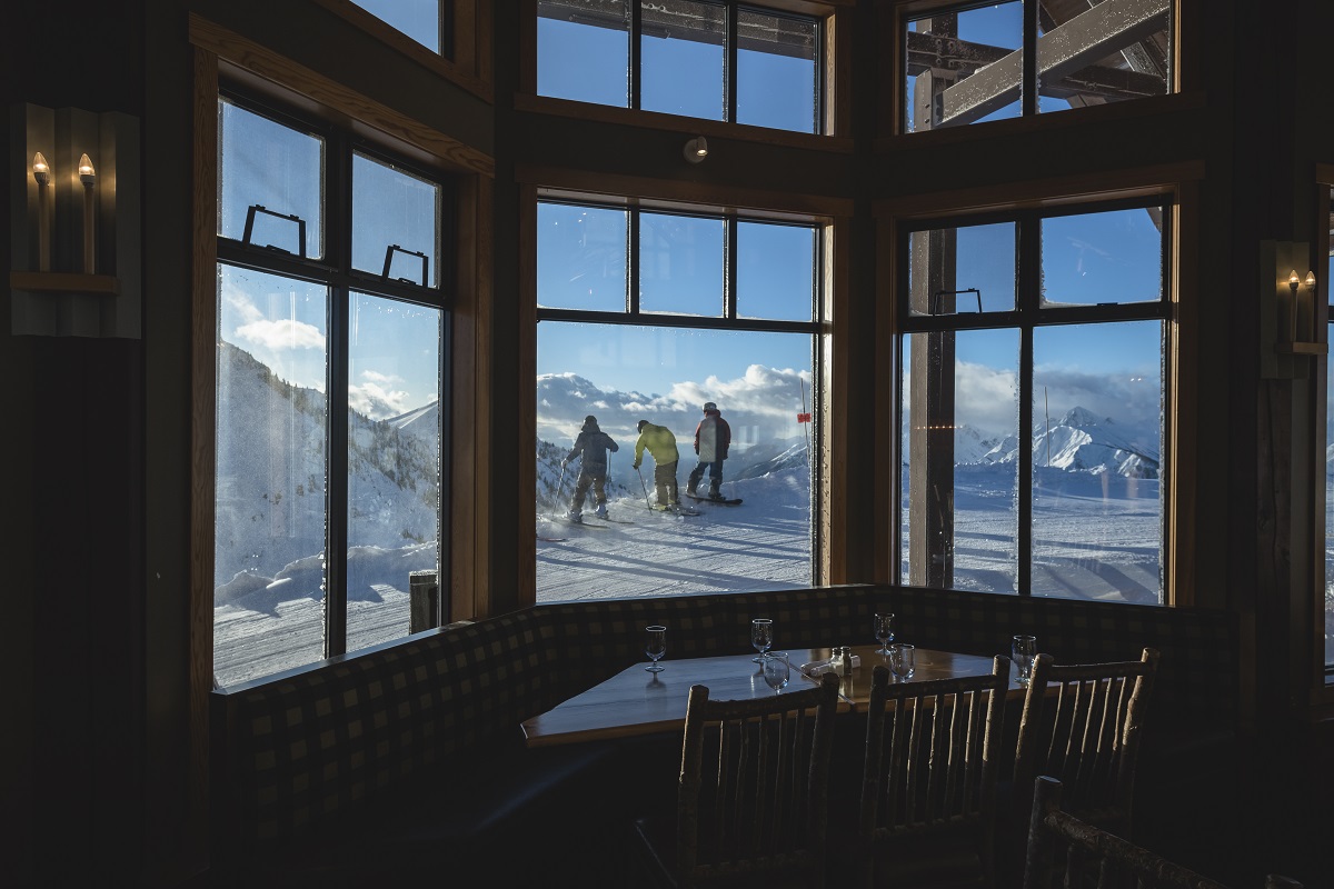 A group of three skiers head out as seen through a window in a restaurant. The restaurant is empty and dark, the ski hill is brightly lit on a blue sky day.
