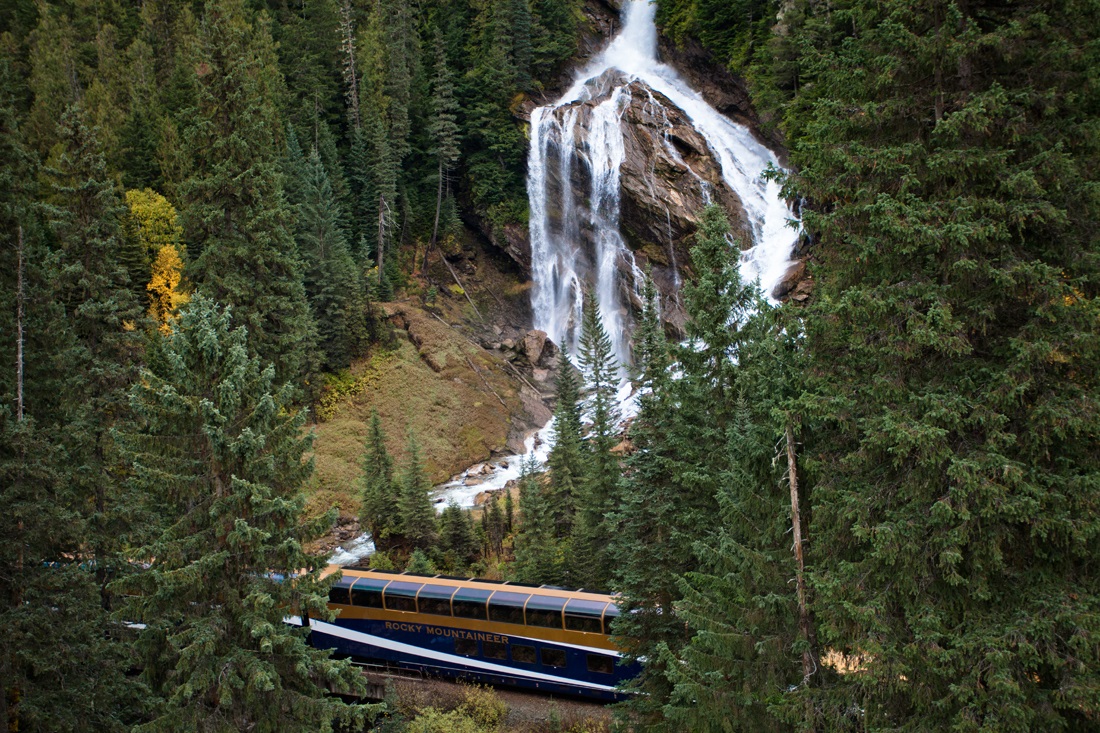 A waterfall down the side of a mountain flows toward a passing train. The waterfall splits near the top with the left side cascading over a ridge and the right curving down the slope.