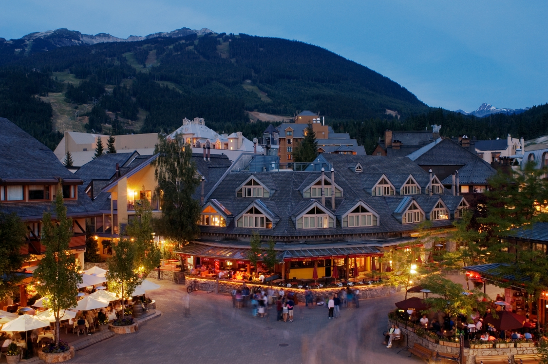 Aerial view of a pedestrian walkway in Whistler Village with hotels and retail space, and forested mountains in the background.