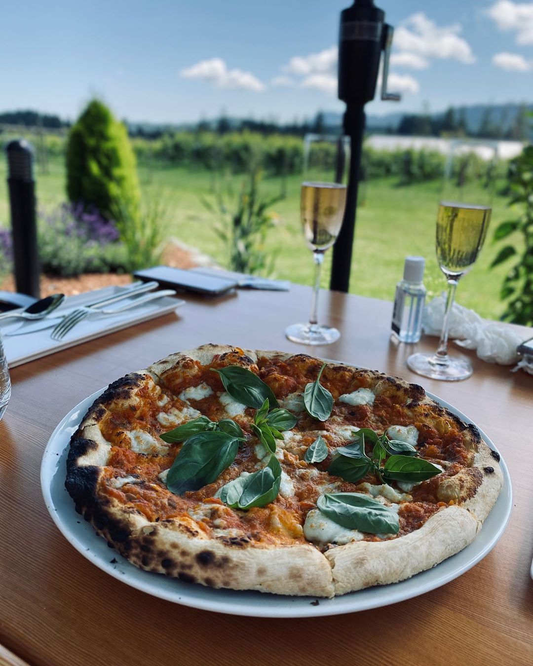 Wood-fired pizza and two glasses of sparkling wine are on a table at Unsworth VIneyards. The table overlooks the vineyard below.
