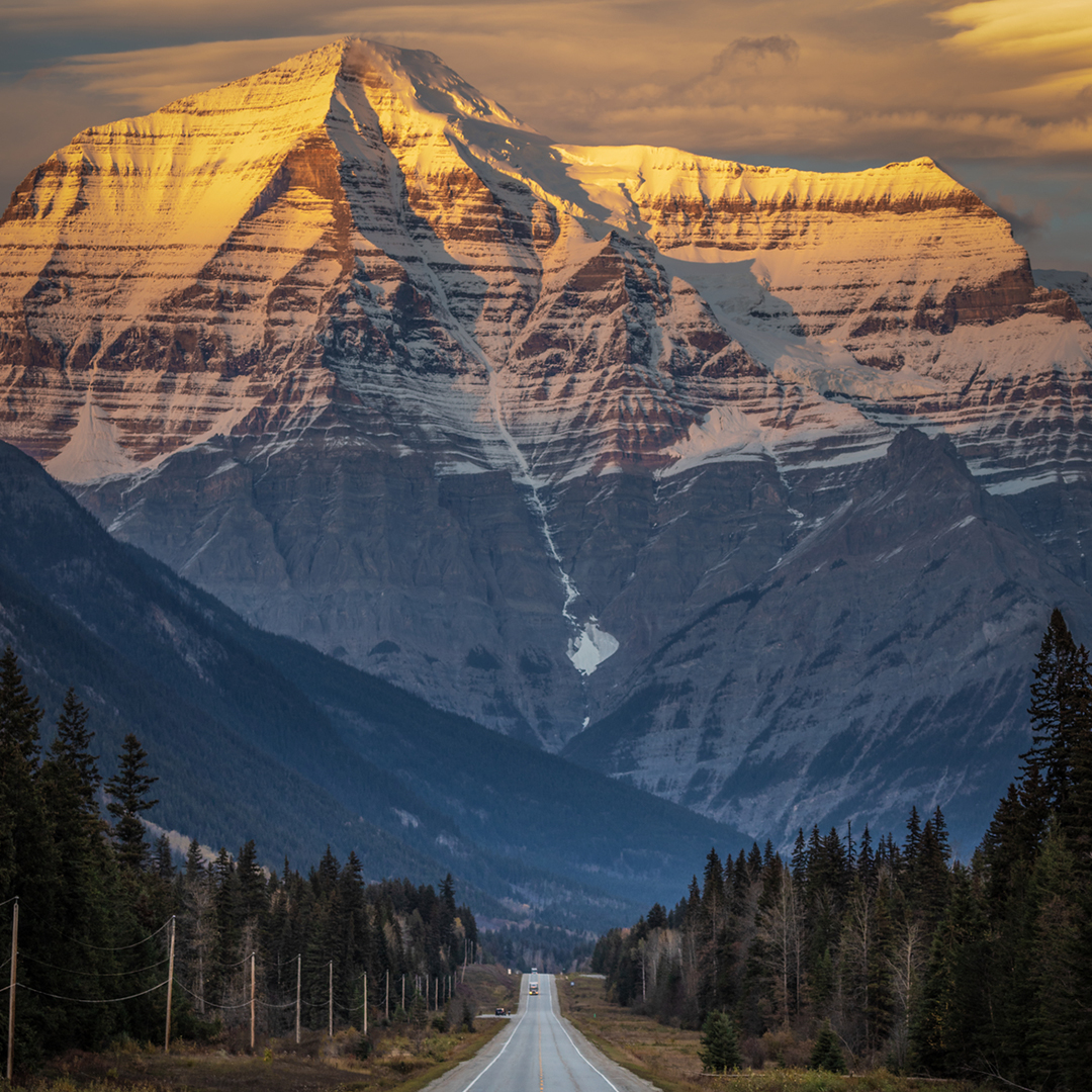 A road runs in the middle of the image with evergreen trees on both side and the dominating Mount Robson as the backdrop. The sunsets over the snowcapped mountain. One vehicle is seen faintly in the distance.