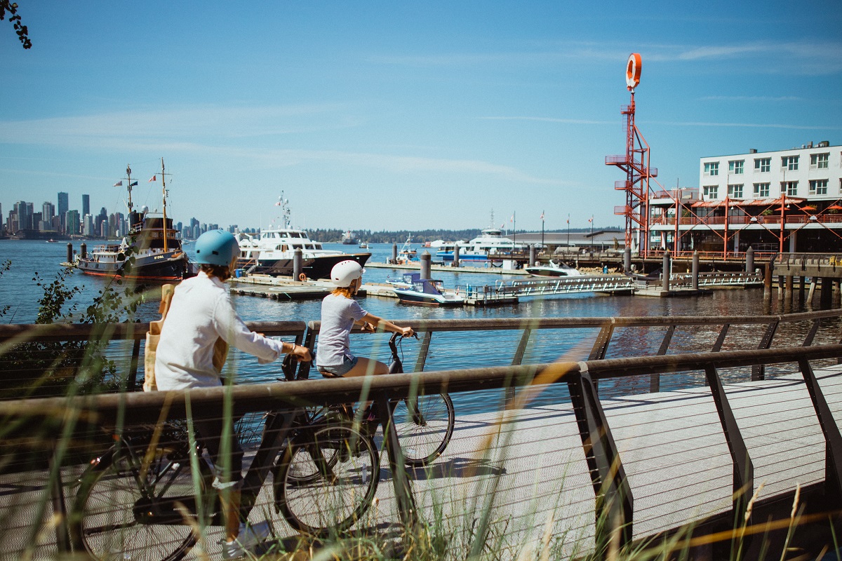 Two cyclists bike across a bridge by the ocean with the Shipyards in the background