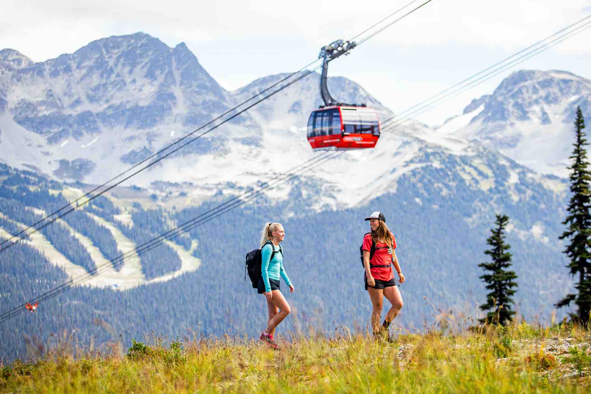 Two women hike in the mountains in the summer carrying backpacks. The red Peak 2 Peak Gondola goes overhead behind them. Whistler mountain is seen behind them.