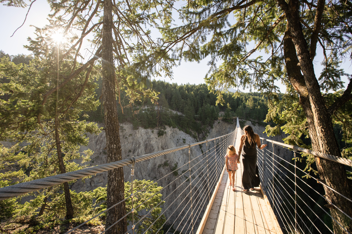 A woman and child hold hands as they walk across the Golden Skybridge (suspension bridge). The woman is wearing a long black summer dress and the child is in shorts and a t-shirt. There are trees they are walking underneath in the foreground.