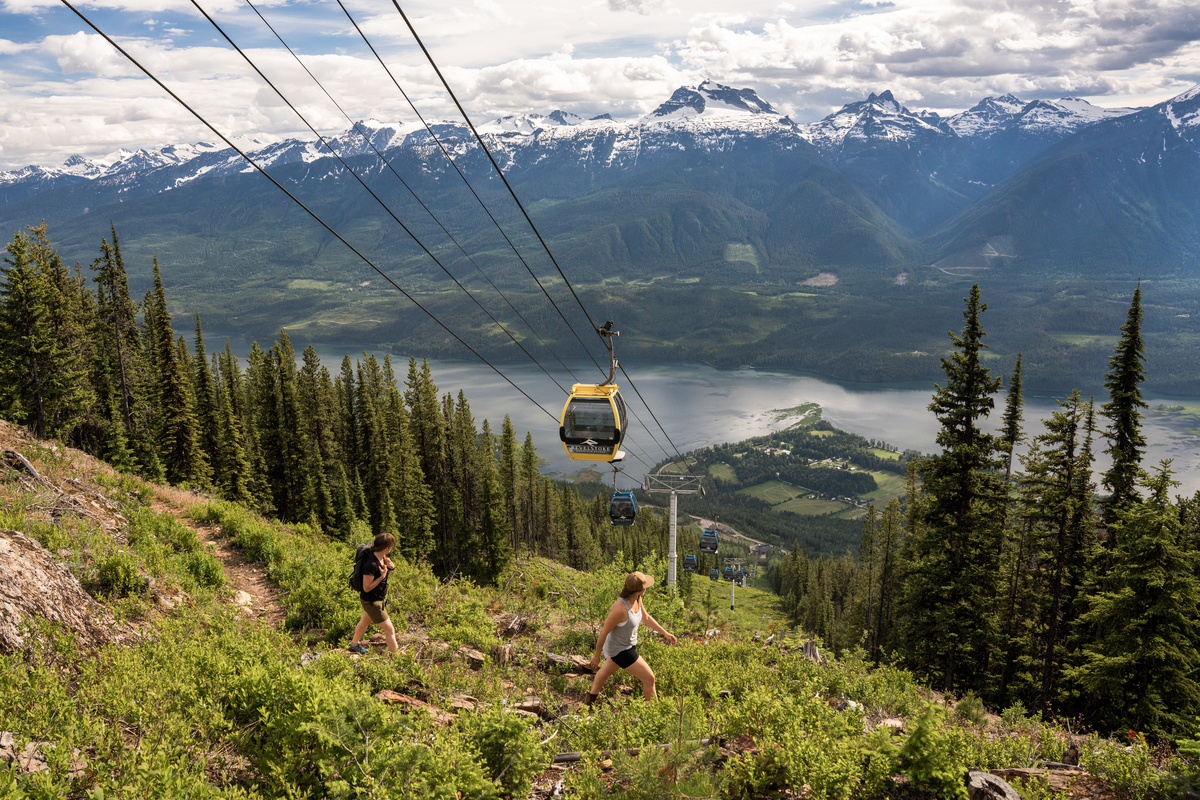 Two hikers walk underneath a gondola going up the mountain in the summertime. There are views of the lake below and snow capped mountain across the lake.