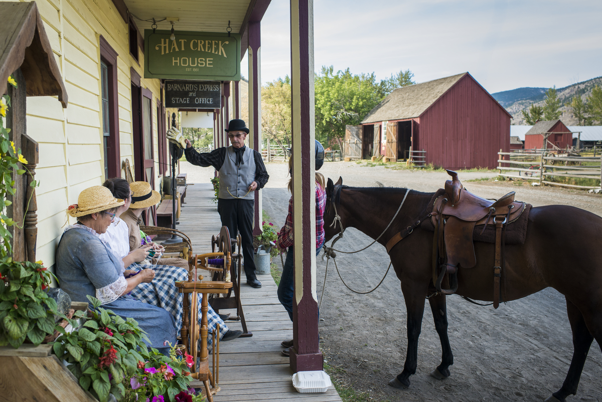 Actors in historical costume sit outside on a bench at Hat Creek Ranch. A horse is tethered to the building.