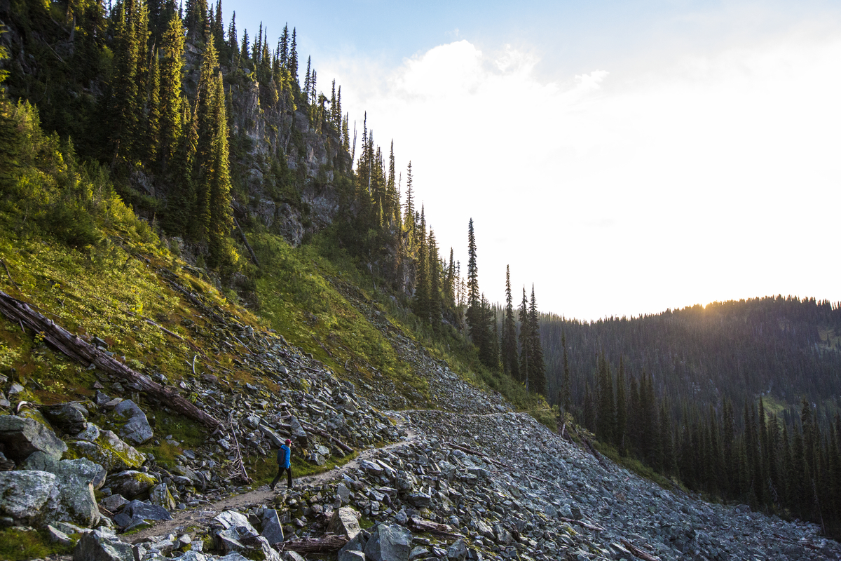 A hiker walks along a trail on the slope of a mountain. There are evergreen trees up on the upward slope. The sun is starting to set behind the mountains in the distance.