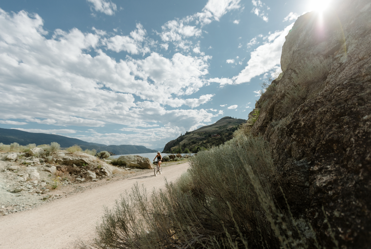 A cyclist on the Okanagan Rail Trail on a sunny day. Partial clouds in the sky. The trail is hard packed dirt and flat next to the lake.