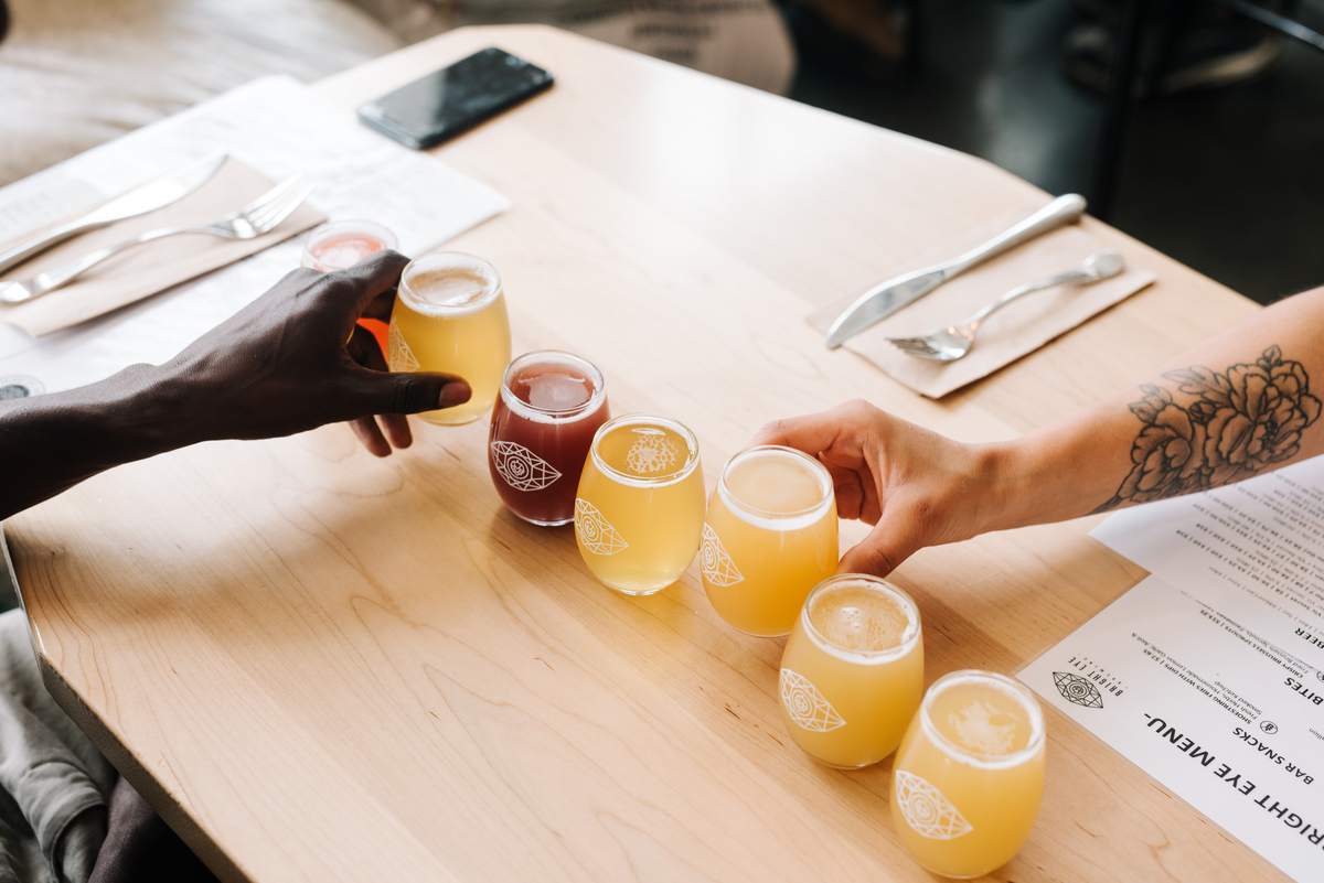 A line up of taster size beers are on a table. Two hands are seen reaching in for a glass.